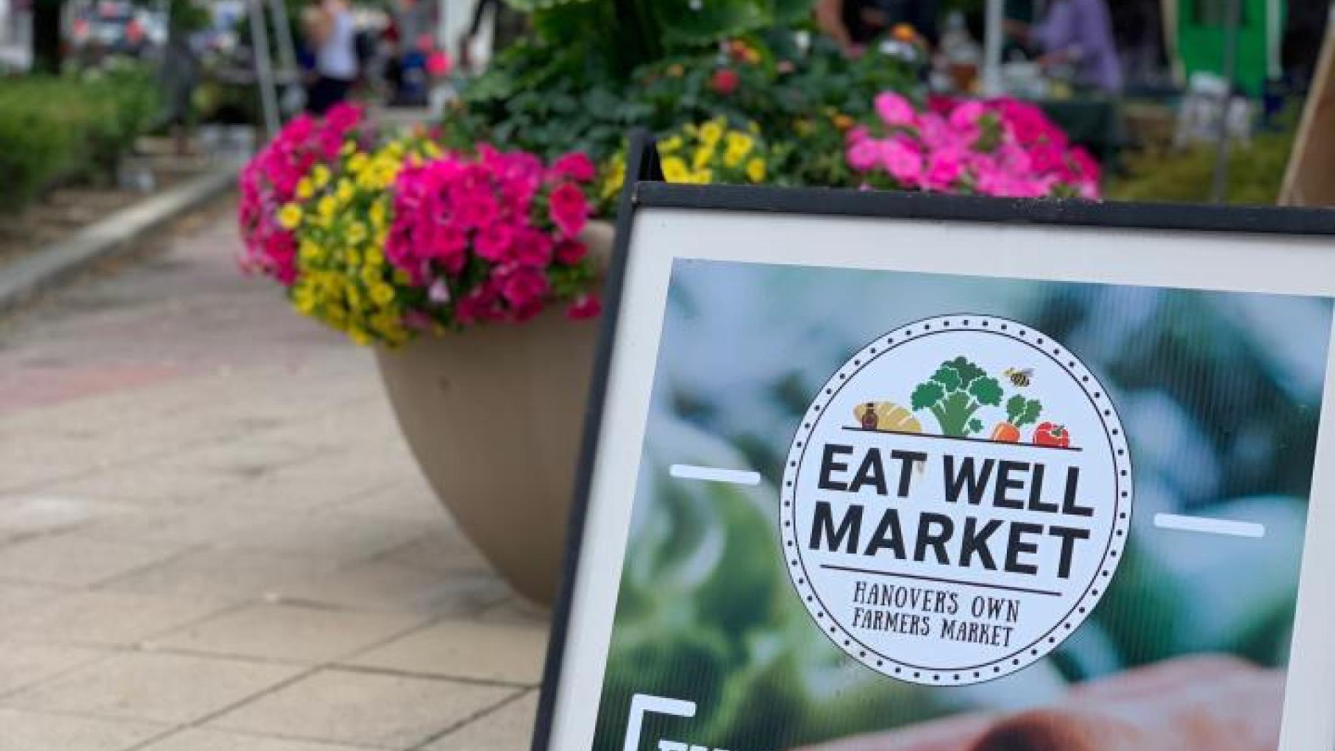 Image of a a frame sign with eat well market every Saturday 9:00 a.m. to 1:00 p.m. eatwellmarket.ca in front of a flower display