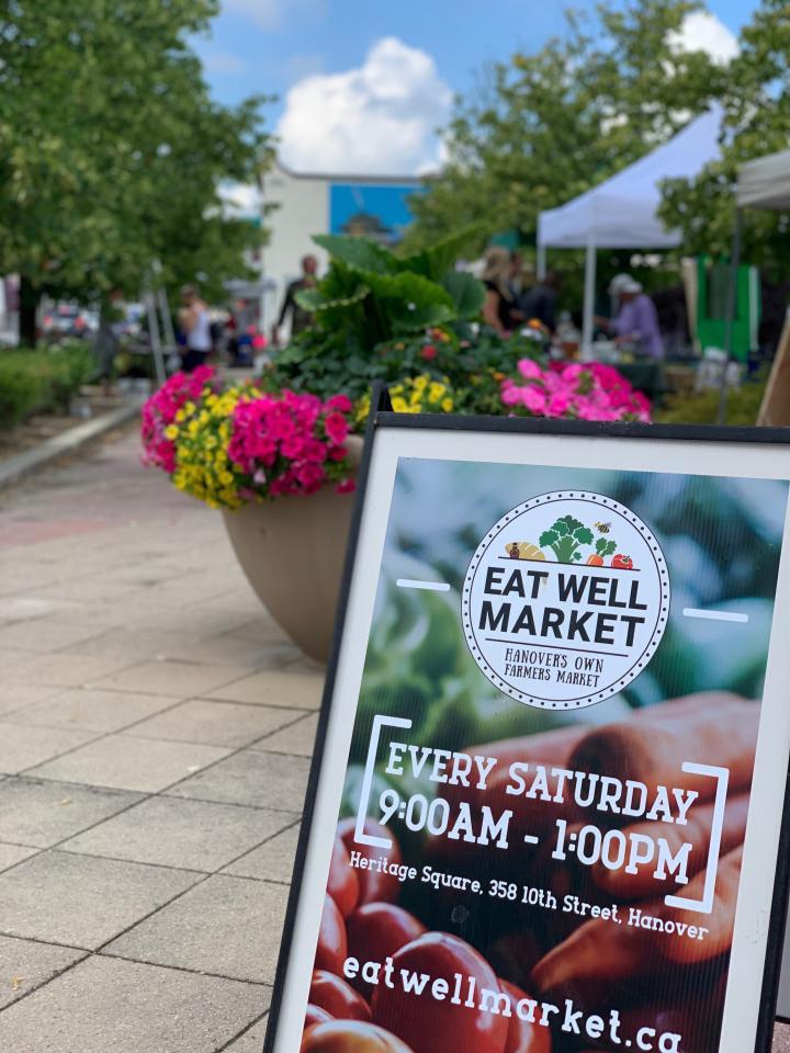 Image of a a frame sign with eat well market every Saturday 9:00 a.m. to 1:00 p.m. eatwellmarket.ca in front of a flower display